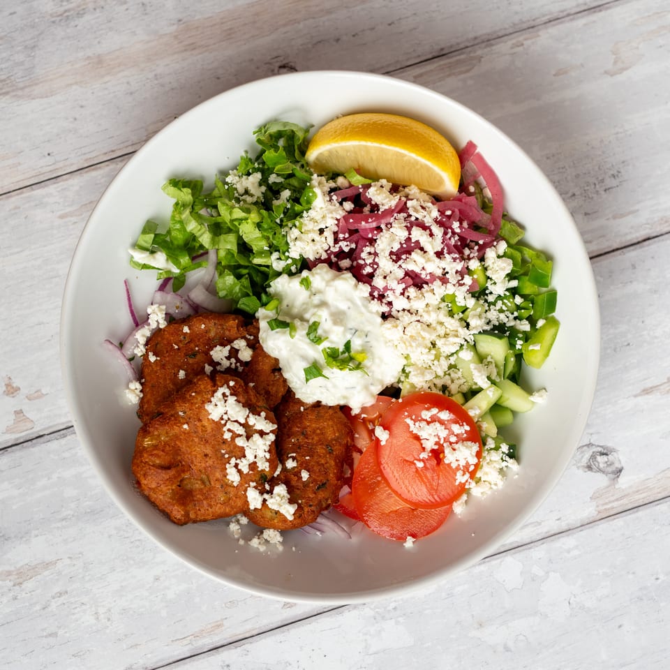 Greek-inspired bowl with falafel, lettuce, cucumber, pickled onions, tomato, tzatziki, feta, and lemon wedge.