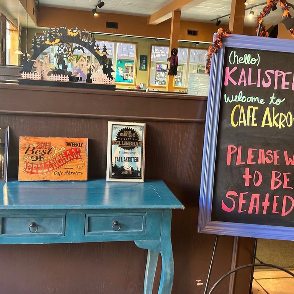 Chalkboard sign welcoming guests to Café Akroteri in Kalispell stands beside a blue console displaying awards and promotional plaques.