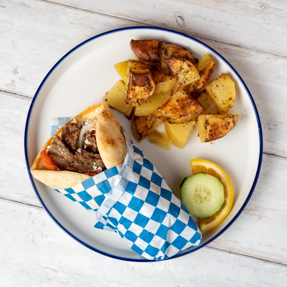 Gyro pita wrapped in blue checkered paper with grilled potatoes, cucumber, and lemon on a plate.