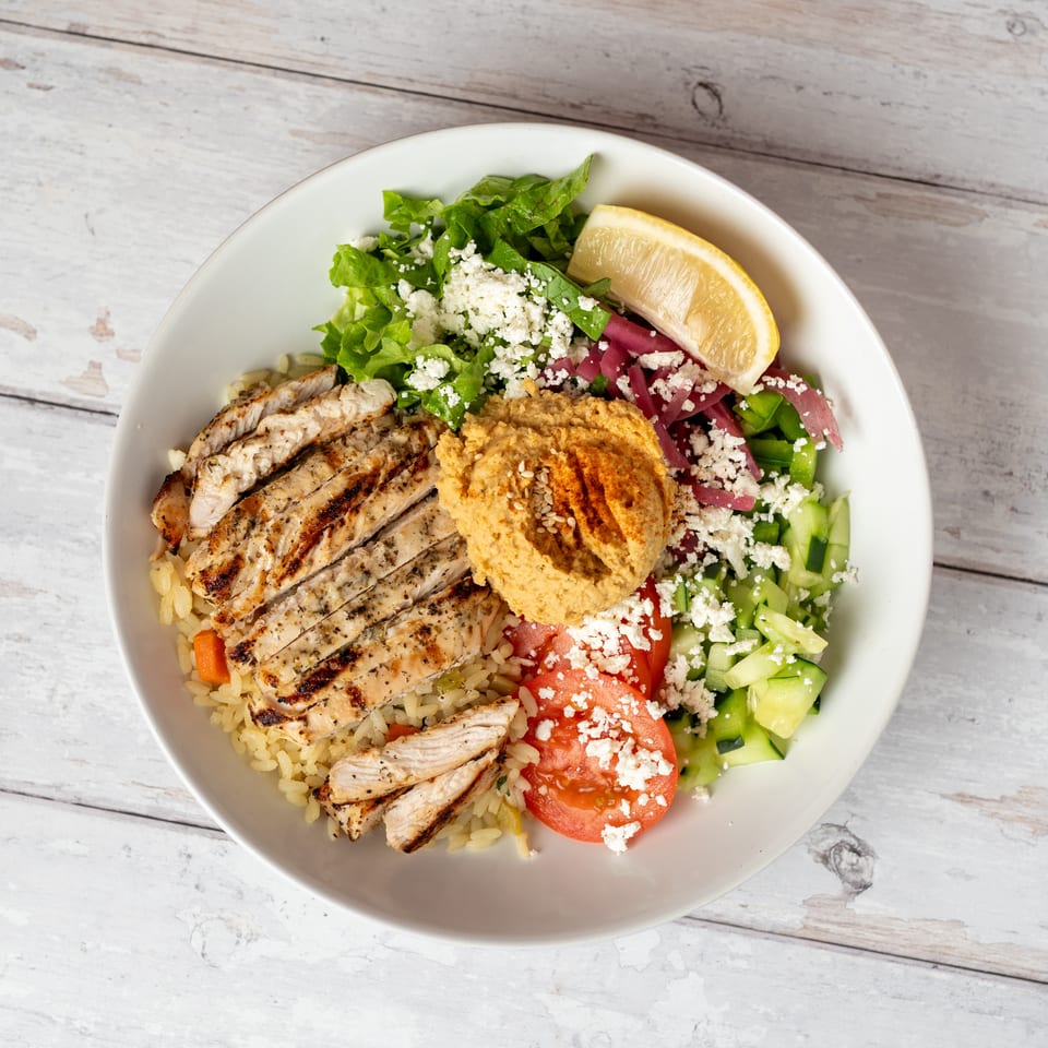 Greek-inspired bowl with grilled chicken, rice, hummus, cucumber, tomato, greens, feta, pickled onions, and lemon.