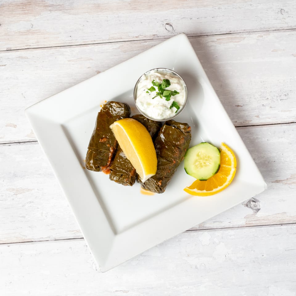 Stuffed grape leaves served with lemon, cucumber, orange slices, and a cup of creamy herb dip on a white plate.