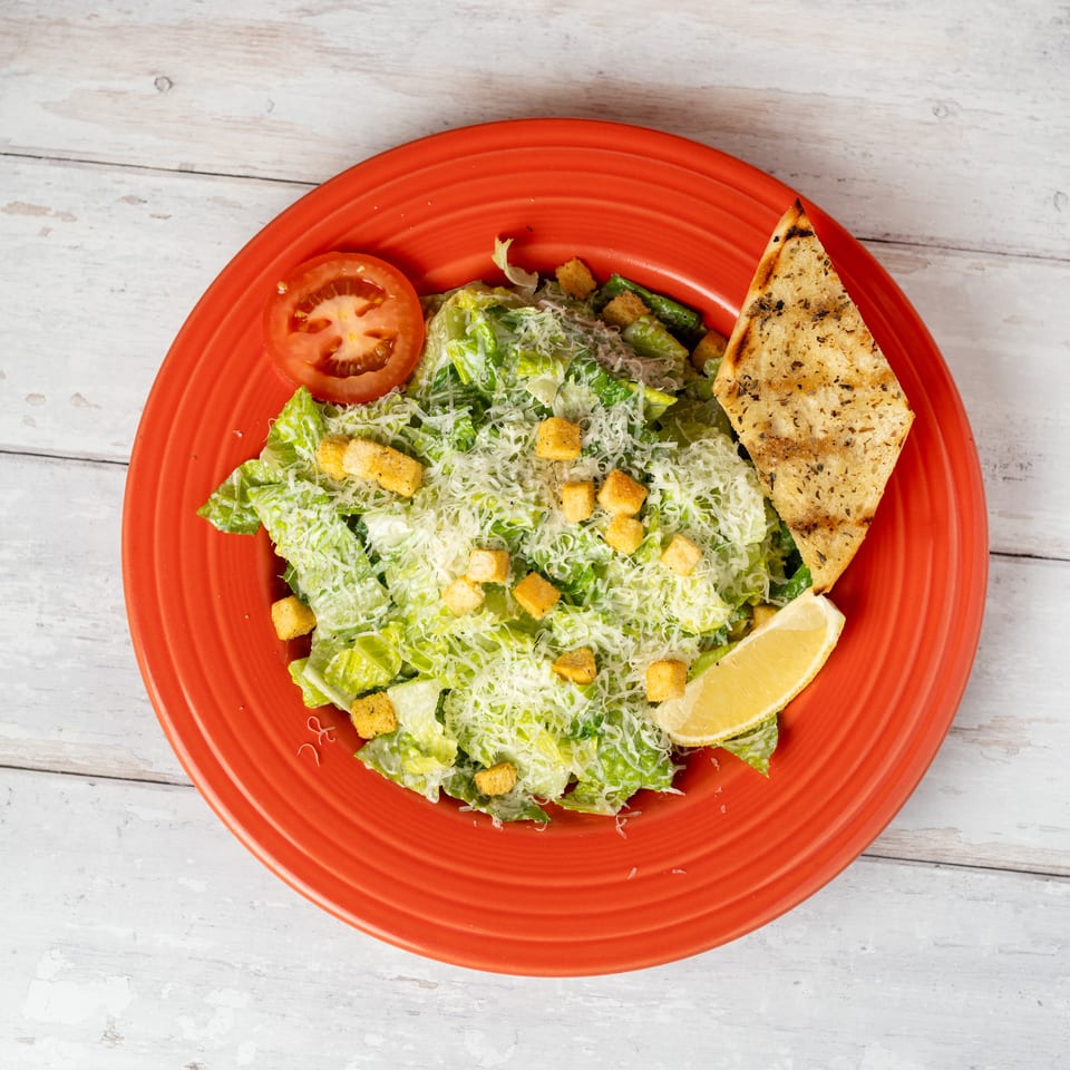 Caesar salad with romaine lettuce, croutons, grated cheese, tomato, lemon wedge, and toasted flatbread on a red plate.