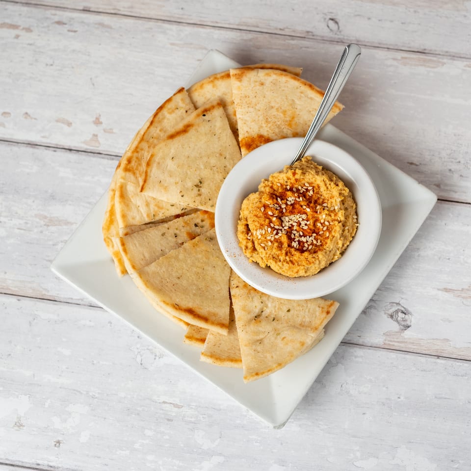 Bowl of hummus topped with paprika and sesame seeds, surrounded by toasted pita wedges on a square plate.