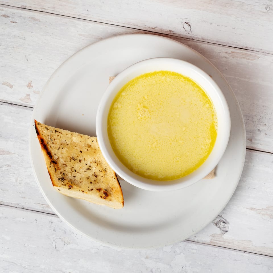 Bowl of creamy yellow soup with toasted herb bread on a white plate atop a rustic white wooden table.