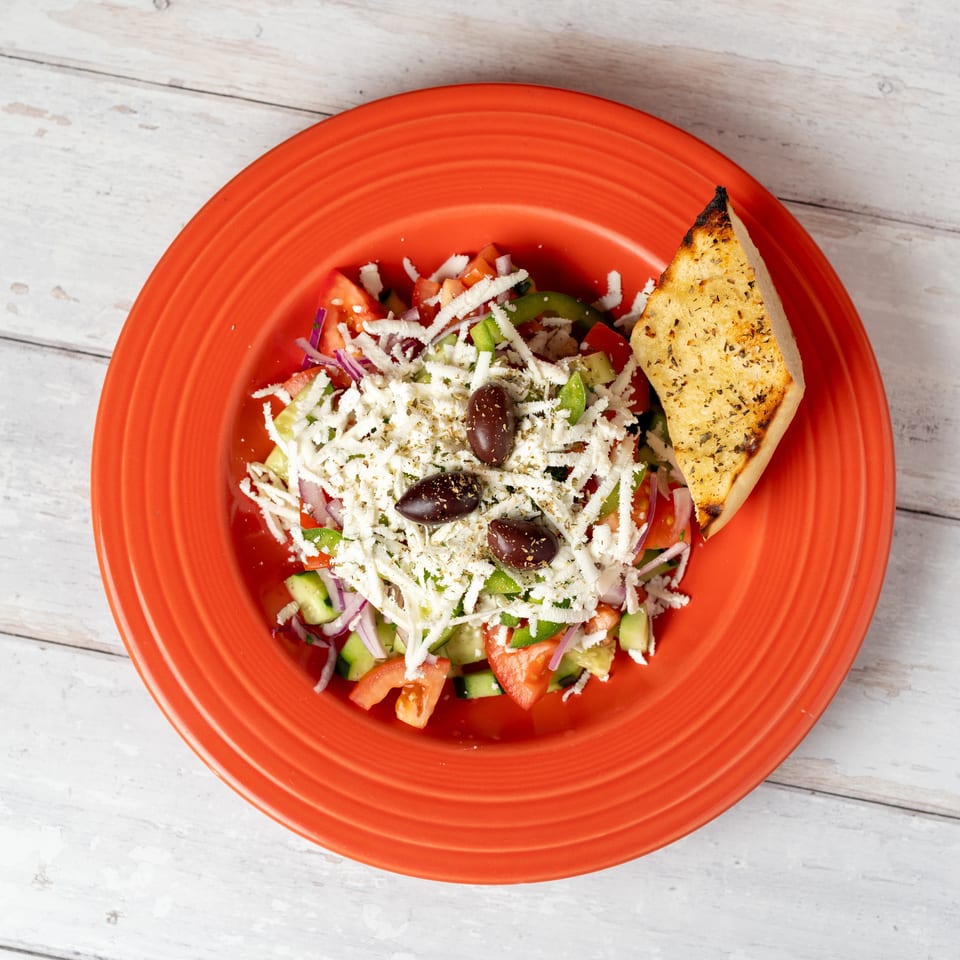 Greek salad with tomatoes, cucumbers, peppers, onions, olives, shredded feta, and toasted pita on a red plate.