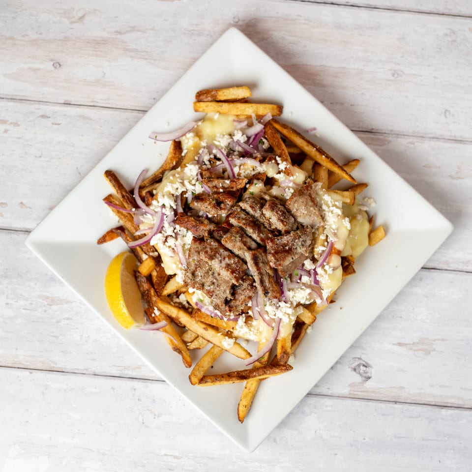 Greek-style loaded fries topped with grilled meat, melted cheese, feta, red onion, and lemon on a square plate.